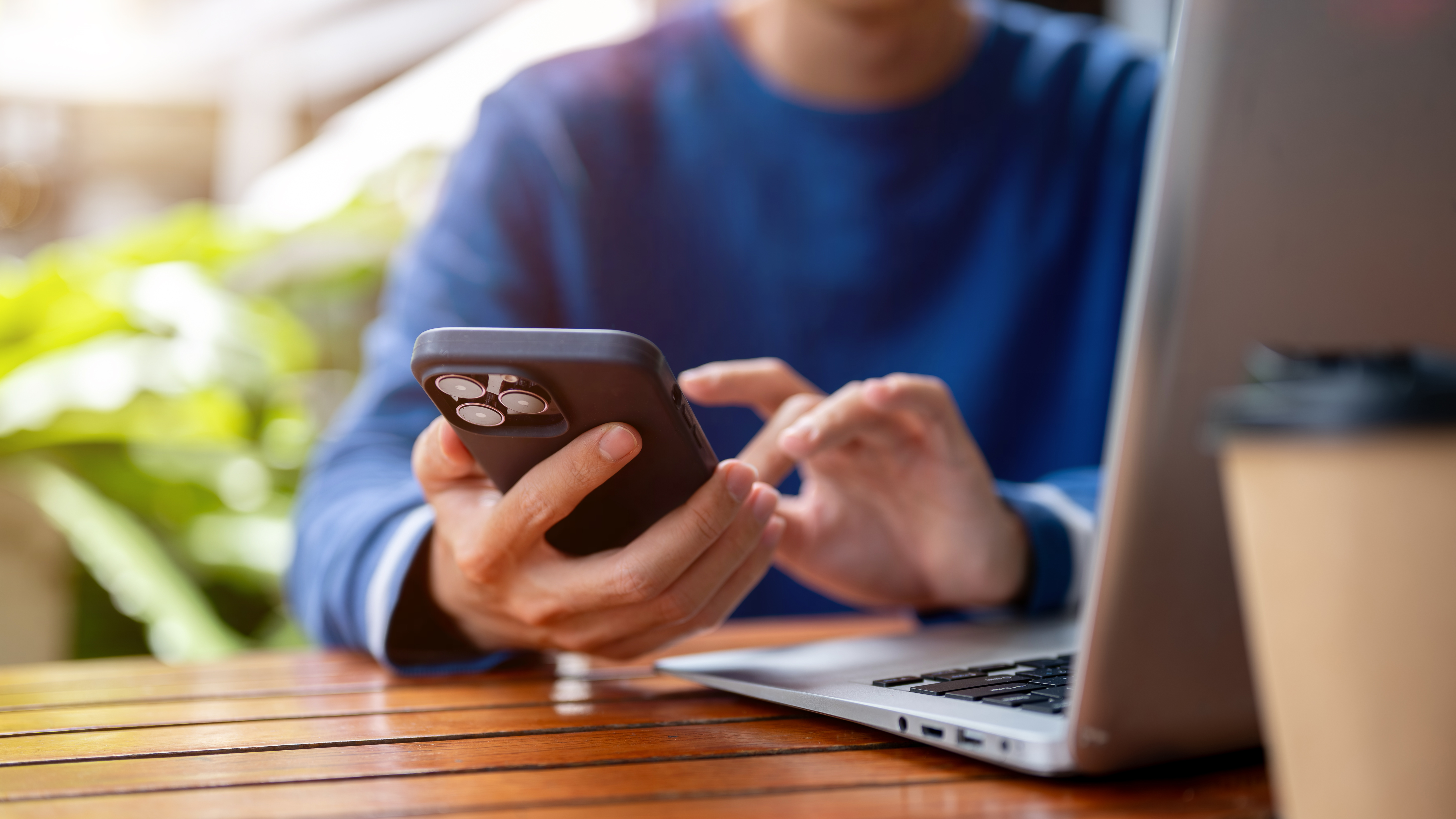 Close up of hands holding and touching on phone aside laptop at wooden table in cafe or canteen. College student, Working or study, Sitting Outdoor.
